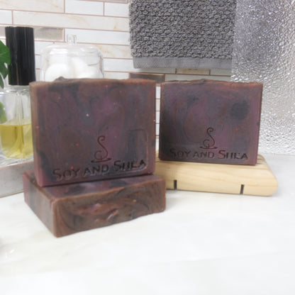 Three rectangular bars of soap are presented on a bathroom countertop next ot a bottle of perfume and a hanging towel.   One soap lies flat on the bench, with another standing upright on it.  To the back right, a third soap stands on a wooden soap rack.  THe soaps have a swirling pattern in dark shades of red and purples.