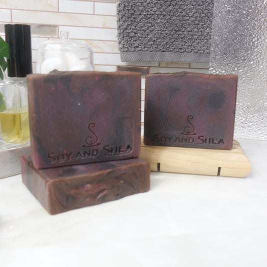 Three rectangular bars of soap are presented on a bathroom countertop next ot a bottle of perfume and a hanging towel.   One soap lies flat on the bench, with another standing upright on it.  To the back right, a third soap stands on a wooden soap rack.  THe soaps have a swirling pattern in dark shades of red and purples.