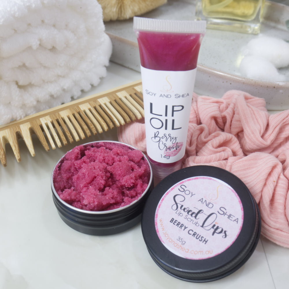 A small black tin with the lid removed revealing a cerise Lip Scrub sits on a marble tile surface next to a small, clear tube of cerise coloured Lip Oil.   This rests againsts a wooden comb and pink scrunchie. 