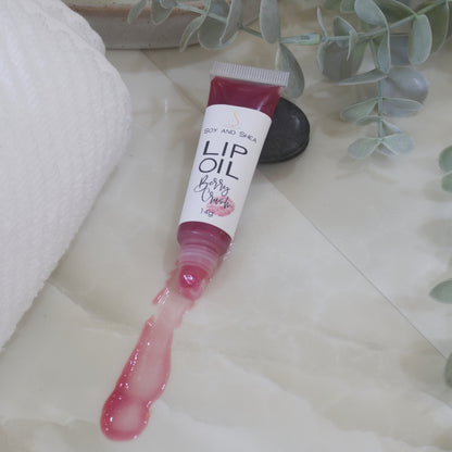 A tube of Lip Oil lies on a marble tile surface next to a white towel and some eucalyptus leaves.   The tube is clear with a white product label and filled with a cerise Lip oil.  The cap is removed and a smear of Lip oil is on the bench showing the translucent effect it has