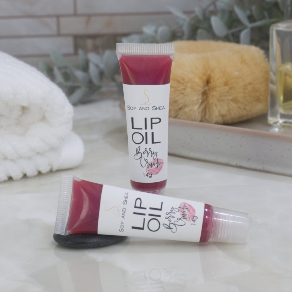 Two tubes of Lip Oil are presented on a bathroom countertop with a blurred background of towels, greenery and body brush.   One tube stands upright while the other lays flat in front of it.  The tubes are clear with a white product lable and are filled with a cerise Lip oil.