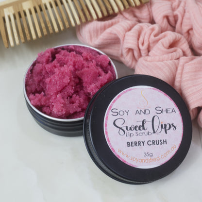 A black tin of Lip Scrub sits on a marble countertop next to a scrunchie and wooden comb.  The lid has been removed and rests on the side of the tin revealing a cerise scrub