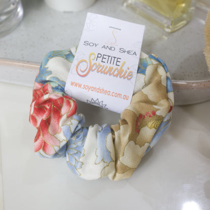 A small scrunchie with label identifying it as Petite, lays resting against a stone tray on a bathroom counter.  The fabric design has large bold flowers in shades of red, blue and gold