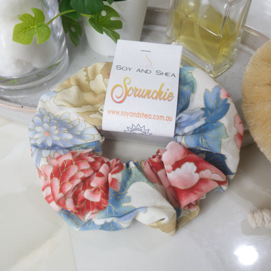 A scrunchie lays on a bathroom counter top surrounded by a perfume bottle, jar of cotton balls and a plant.  The scrunchie features a bold floral pattern in shades of red, blue and gold.