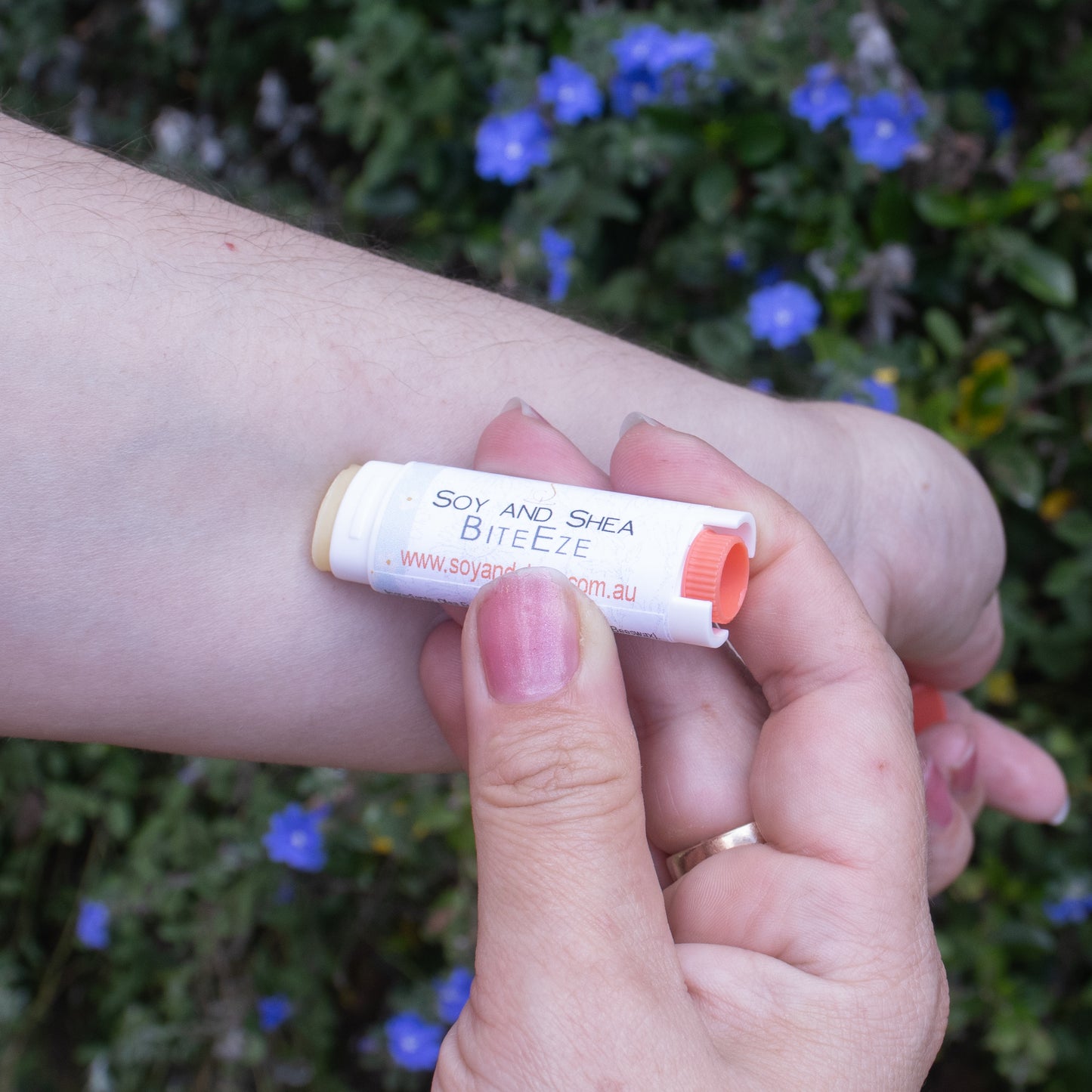 A person applies BiteEze Balm from Soy and Shea, which is infused with Eucalyptus essential oil, to their arm. The background is adorned with green leaves and small blue flowers. The person's fingernails are painted light pink, and they are wearing a gold ring on their ring finger.