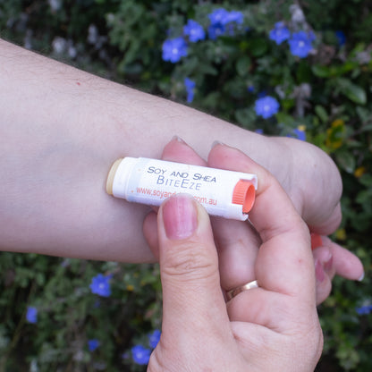 A person applies BiteEze Balm from Soy and Shea, which is infused with Eucalyptus essential oil, to their arm. The background is adorned with green leaves and small blue flowers. The person's fingernails are painted light pink, and they are wearing a gold ring on their ring finger.