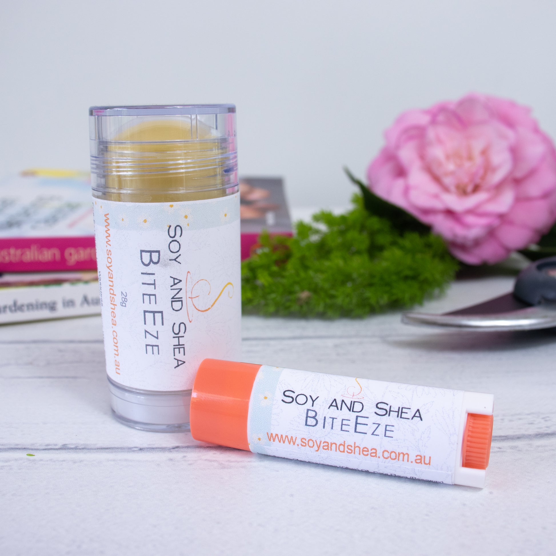 Two tubes of Soy and Shea's BiteEze Balm are displayed on a white surface. One is in a clear applicator standing upright while the other is in a smaller orange-capped container lying down. The background features a pink flower, greenery, and books. The soothing formula includes eucalyptus essential oil for added relief.