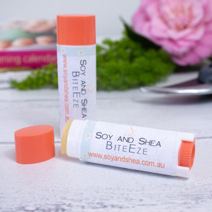 A close-up of two cylindrical tubes of BiteEze Balm from Soy and Shea with orange caps on a white surface. One tube stands upright with its cap off, revealing the balm infused with lavender essential oil, while the other lies down with its cap on. Blurred flowers and foliage are visible in the background.