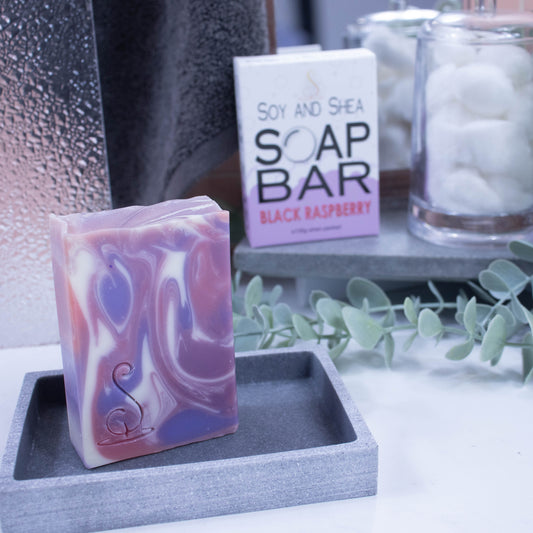A tall rectangular bar of soap sits on a grey soap dish. The soap features swirls of colour in white, pink and purple. Behind the soap is a shower screen and towel along with a box the soap was presented in. The box has a white top with pale mauve bubble border.