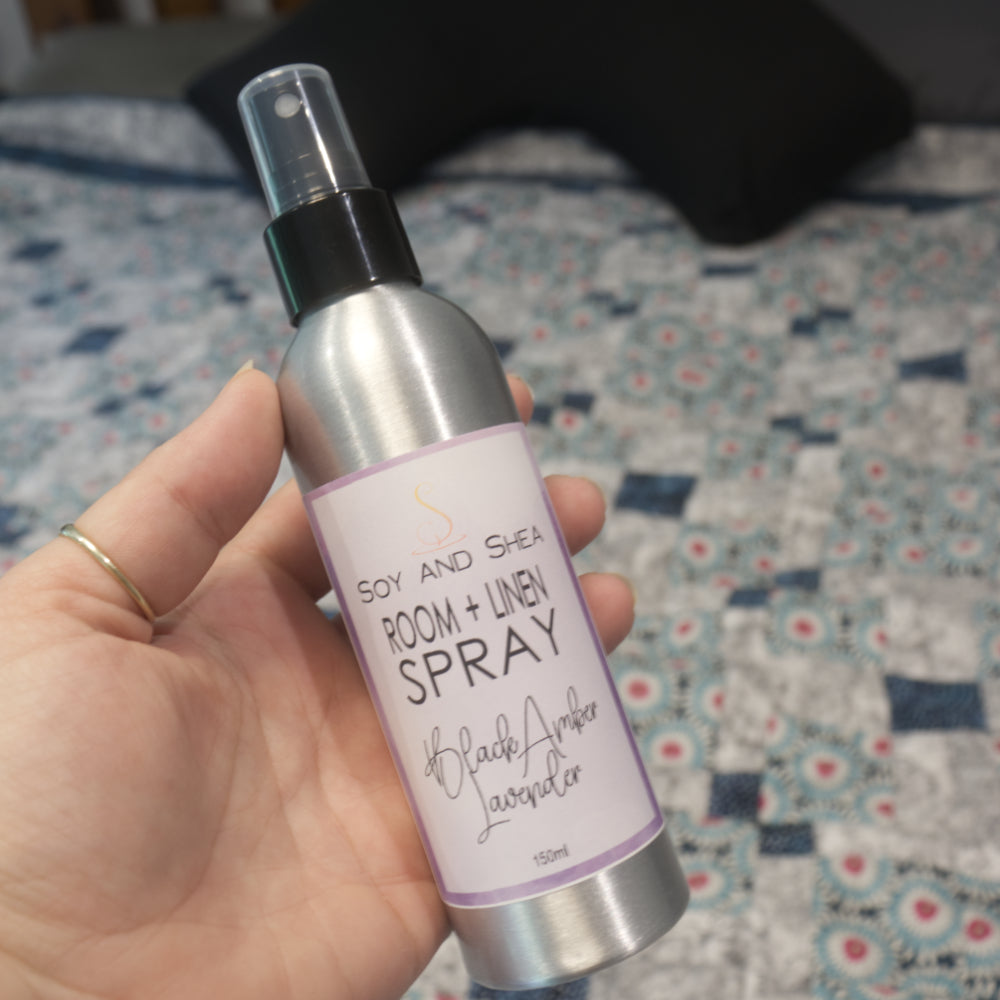 A hand holds an aluminium bottle with black spray cap agains a blurred background of a blurred bed with patchwork quilt and pillows.   The bottle features a decorative product label naming it as Room + Linen Spray and scented in Black Amber Lavender.