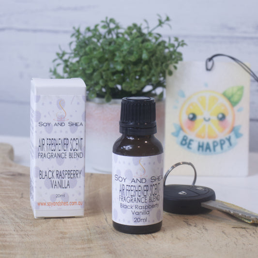 A small amber bottle with black lid labelled AIR FRESHENER SCENT stands on a wooden surface next to the box packaging.  In the background is a car key, air freshener and greenery.
