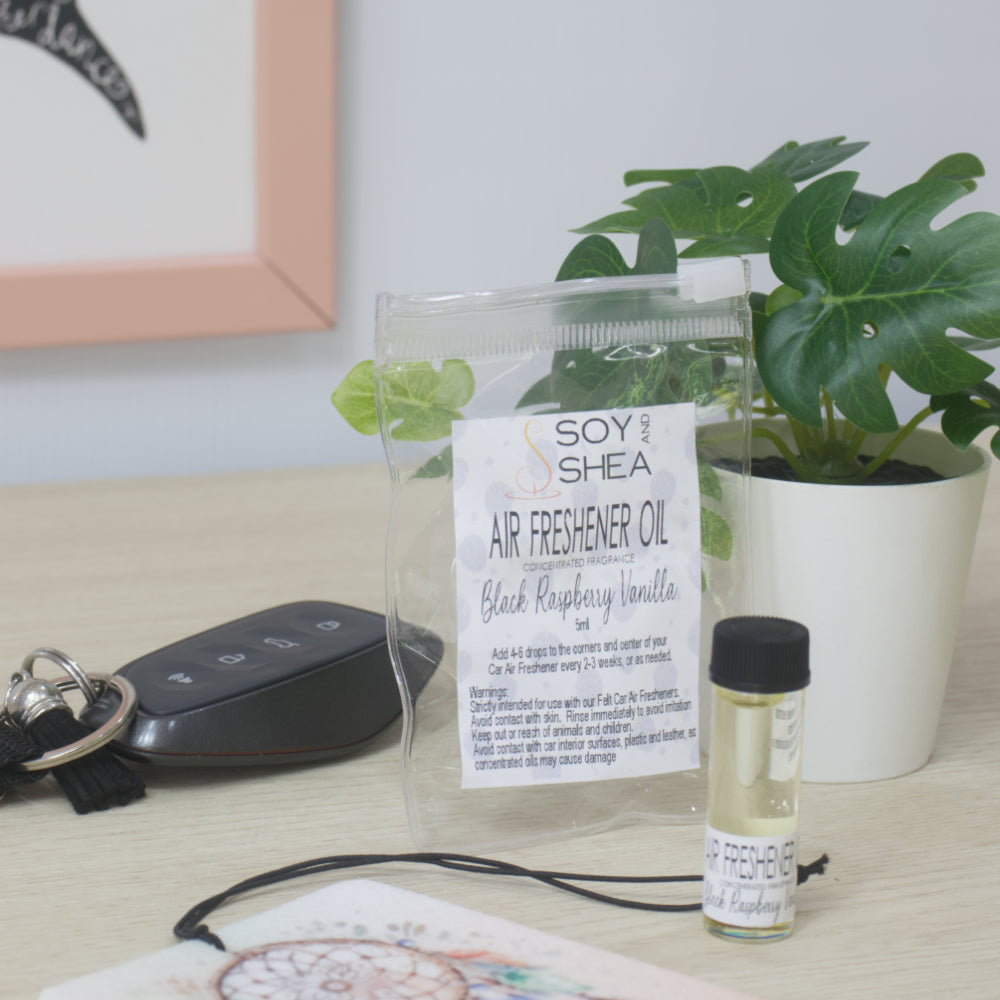 A small clear bottle with black cap of Soy's Shea Air Freshener Oil next to a clear resealable pouch, displayed on a light wood tabletop with a car key, a felt air freshener, and a potted plant in the background.  