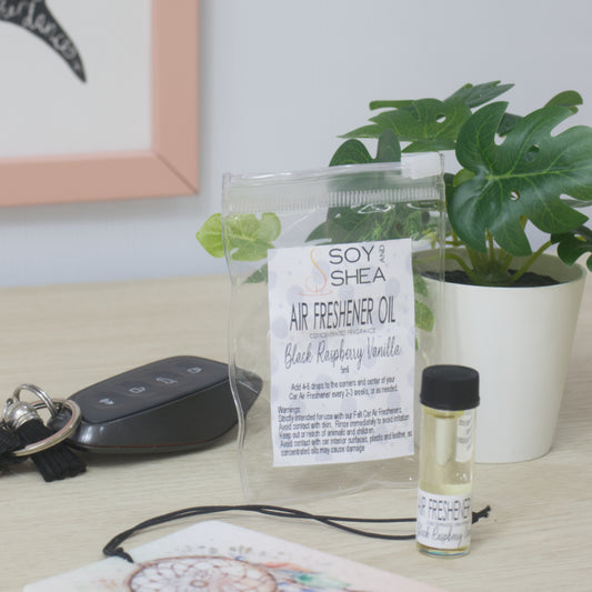 A small clear bottle with black cap of Soy's Shea Air Freshener Oil next to a clear resealable pouch, displayed on a light wood tabletop with a car key, a felt air freshener, and a potted plant in the background.  