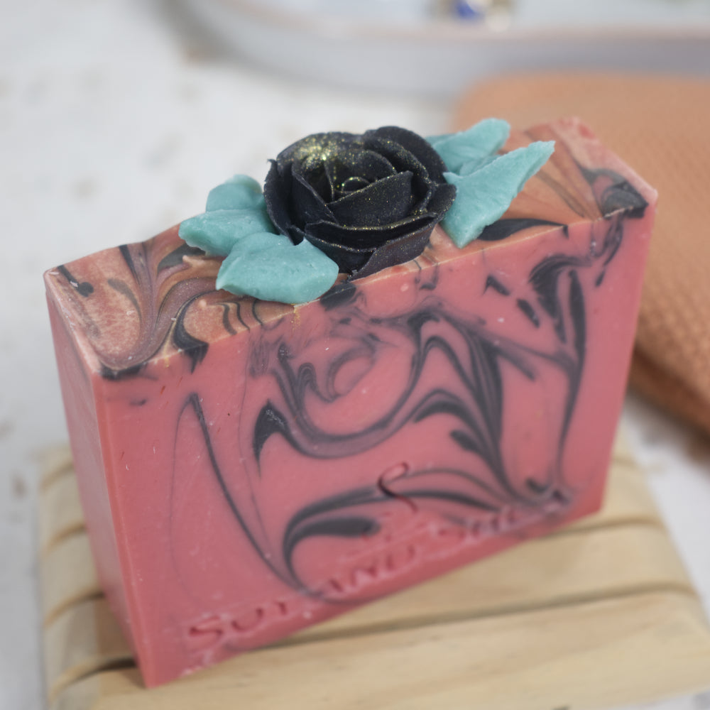 A red bar of soap with black swirls sits on a wooden soap dish with a blurred background.  The  image shows a close up of the soap top that features a black rose piped with soap and surrounded by green leaves