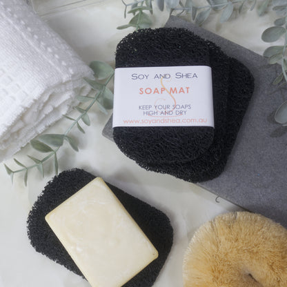 A flatlay image showing a collection of Black Soap mats, displayed on a bathroom countertop along with a white towel, body brush and greenery.  Three soap mats are stacked upon each other in a fan shape on a grey stone tray with the top one featuring a product label.  A fourth mat lies to the bottom of the image with a cream soap lying on it.