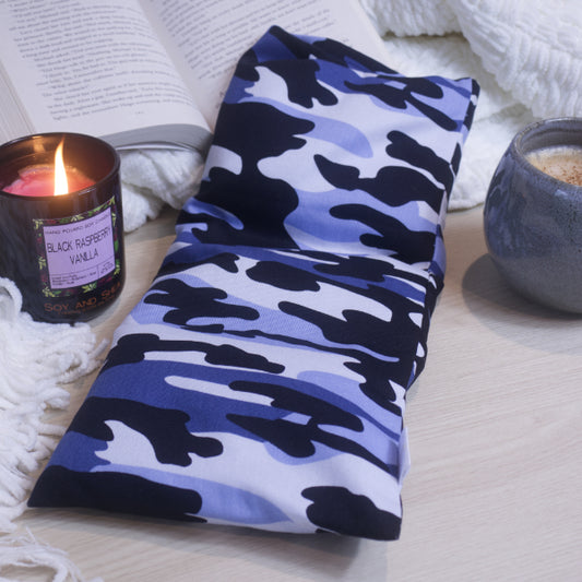 A blue and black camouflage heat pack placed on a table with a lit candle and a cup of coffee sits in the background.  The heat pack is rectangular in shape