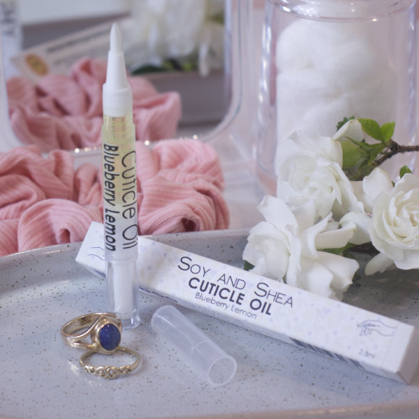 A clear, narrow tube with the lid removed showing a brush tip stands upright on a stone tray.  Beside it sits a cardboard box labelled CUTICLE OIL and a set of gold rings.  In the background is a bathroom counter top with a pink scrunchie, small mirror, jar of cotton balls and a some white  flowers.