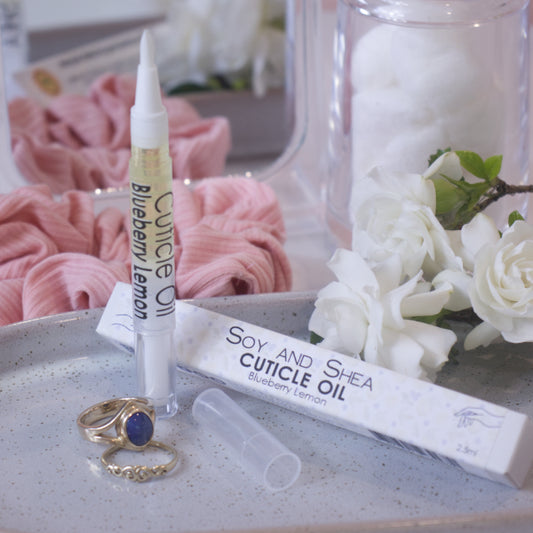 A clear, narrow tube with the lid removed showing a brush tip stands upright on a stone tray.  Beside it sits a cardboard box labelled CUTICLE OIL and a set of gold rings.  In the background is a bathroom counter top with a pink scrunchie, small mirror, jar of cotton balls and a some white  flowers.