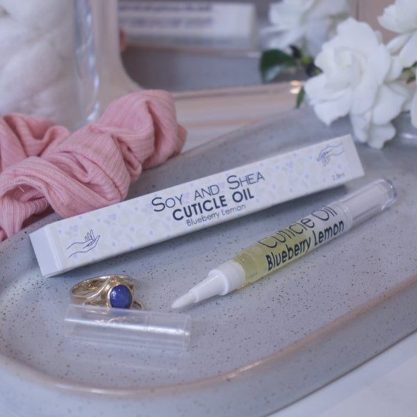 A clear, narrow tube with the lid removed showing a brush tip lies on a stone tray beside a cardboard box labelled CUTICLE OIL and a set of gold rings.  In the background is a bathroom counter top with a pink scrunchie, small mirror, and a some white  flowers.