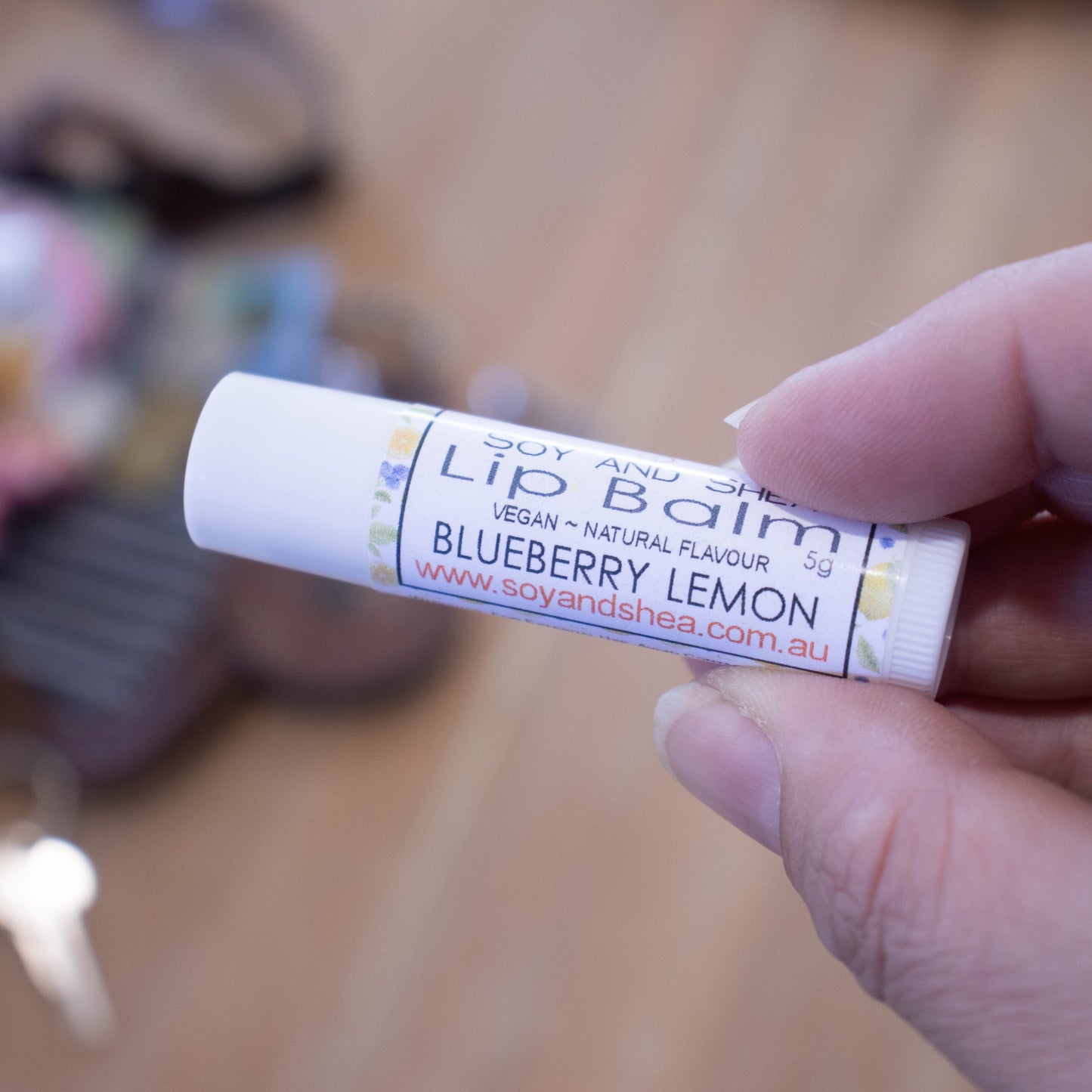 A hand holds a white tube labelled lip balm over a blurred image of an open handbag with contents spilling onto the table.  The lip balms features a white label with watercolour images of blueberrie and lemons