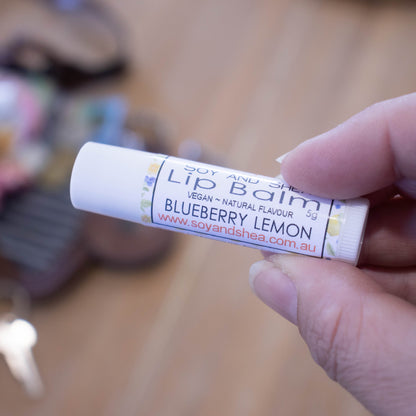 A hand holds a white tube labelled lip balm over a blurred image of an open handbag with contents spilling onto the table.  The lip balms features a white label with watercolour images of blueberrie and lemons