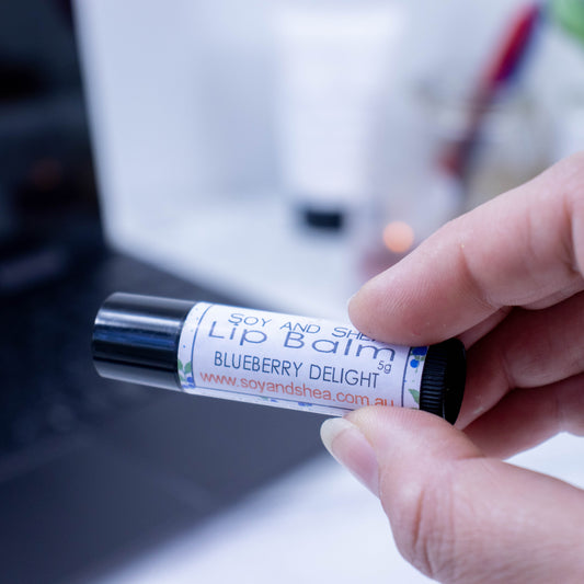 A hand holds a black tube of lip balm against a blurred background with an open laptop computer on a white surface.  The label has text LIP BALM BLUEBERRY DELIGHT on a white background with a border of small blueberries with green leaves.