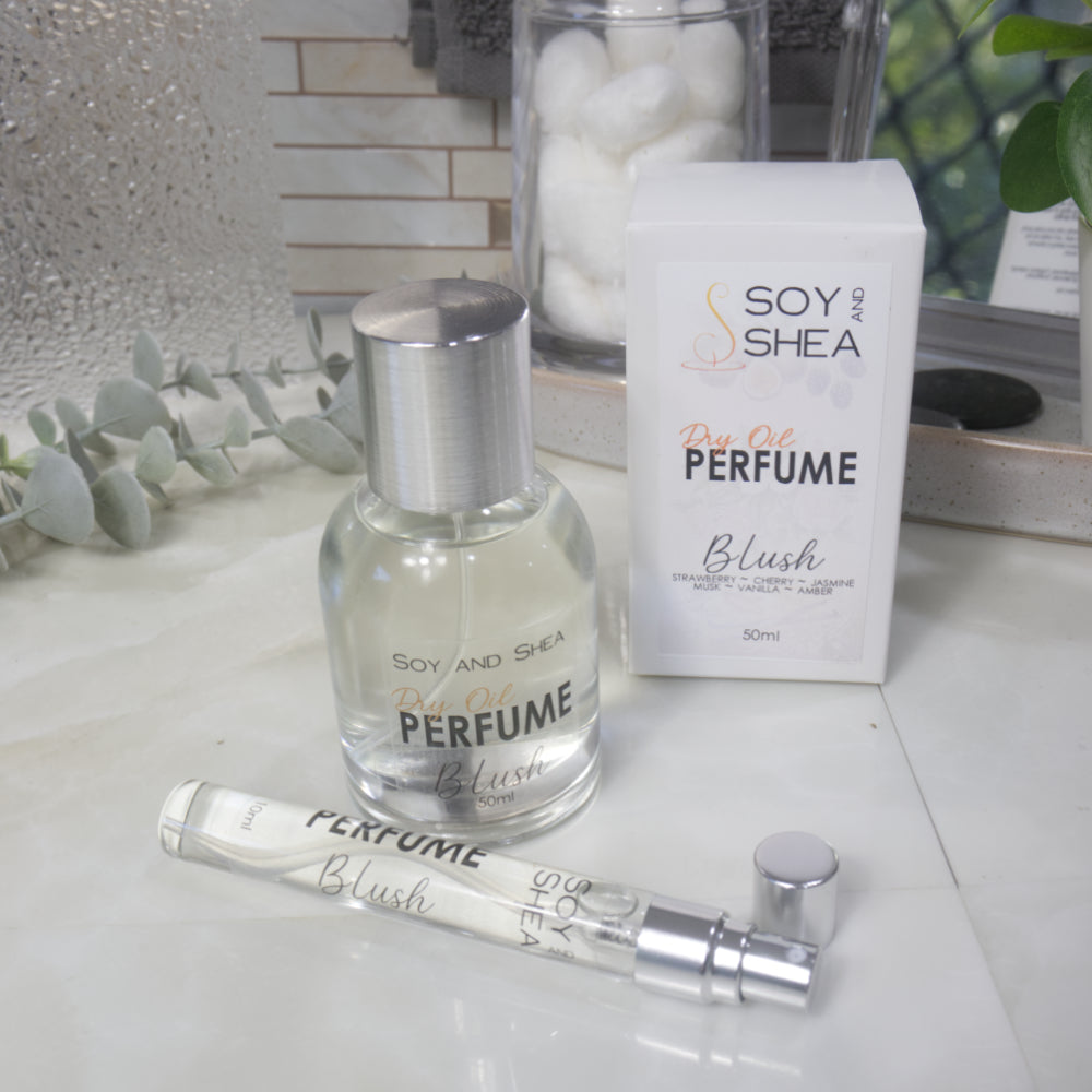 A selection of perfume bottles are presented on a bathroom countertop.  To the front is a clear, narrow, tall bottle labeled as 10ml with a silver cap and behind is a clear round, short bottle with silver lid labelled as 50ml.  Behind sits a white box with label identifying as Dry Oil Perfume Blush.
