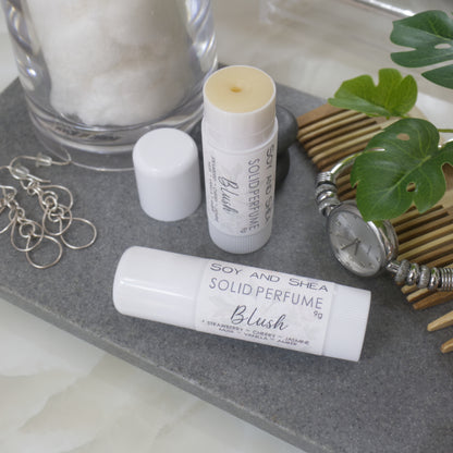Two white tubes of Solid Perfume are displayed on a grey stone tray along with silver earrings, watch and greenery.   One tube lays flat  displaying the fragrance notes while another tube stands upright behind it with the lid removed and to the left revealing a beige balm