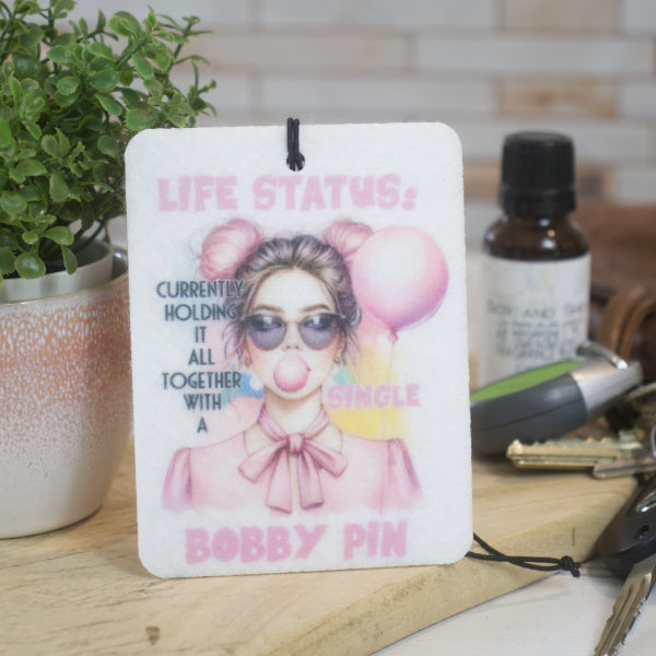 A white car air freshener with a design featuring a cartoon girl holding a pink ballon and blowing pink bubbleum, and text that reads 'LIFE STATUS: CURRENTLY HOLDING IT ALL TOGETHER WITH A SINGLE BOBBY PIN' rests against a pot plant. To the left is a bottle of fragrance and a set of car keys