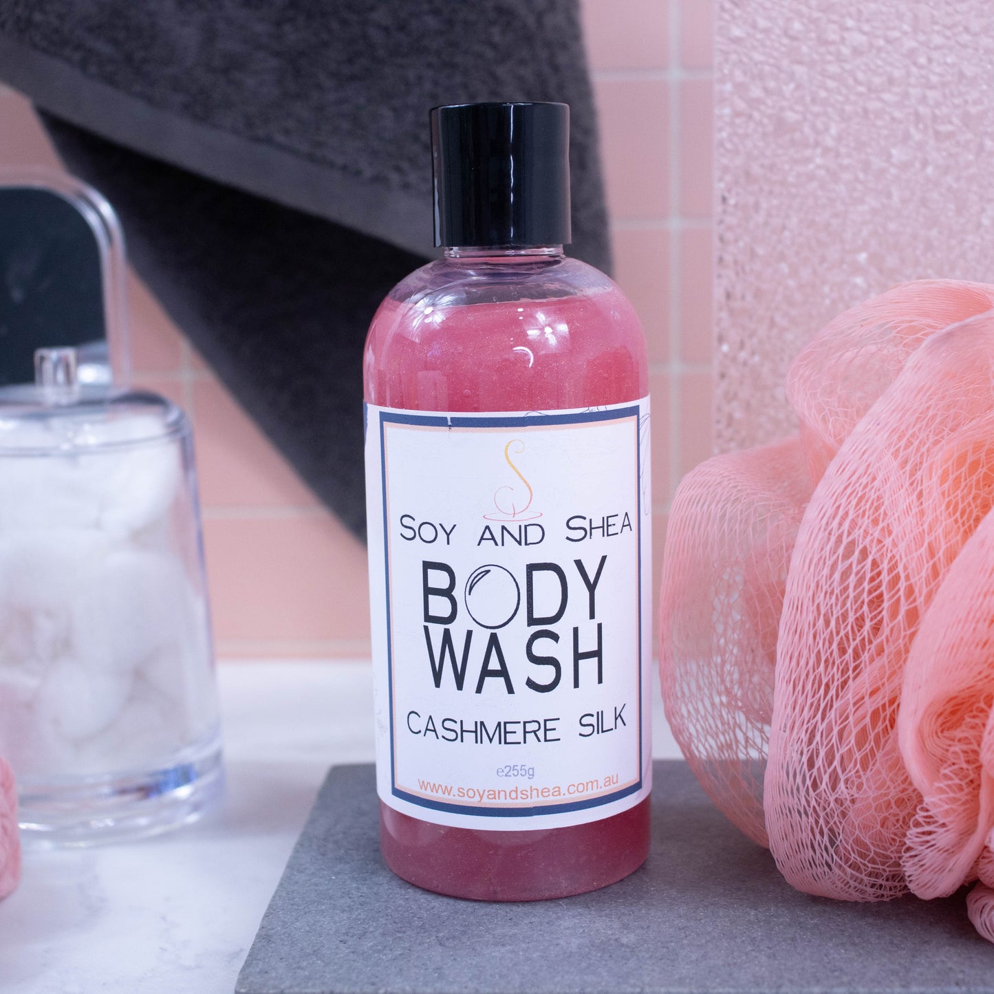 A bottle of Body wash sits on a stone bench.  It is a dark pink liquid and the bottle has a disc cap.  Next to the bottle is a shower sponge  and a towel handing in the background