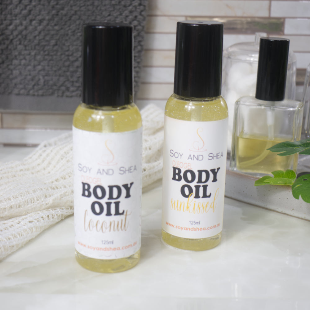 Two clear bottles with black caps sit on a bathroom countertop.  The bottles are clear filled with a golden liquid.  Product labels identify these as an Oleogel Body Oil in two different scents