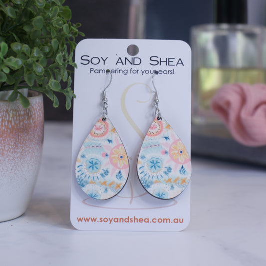 A pair of earrings on silver hooks hang on a white card branded with SOY AND SHEA leaning against a pot with a green leafy plant and a blurred background.  The earrings are teardrop shaped with a boho floral pattern in pale but bright shades of blue and pink