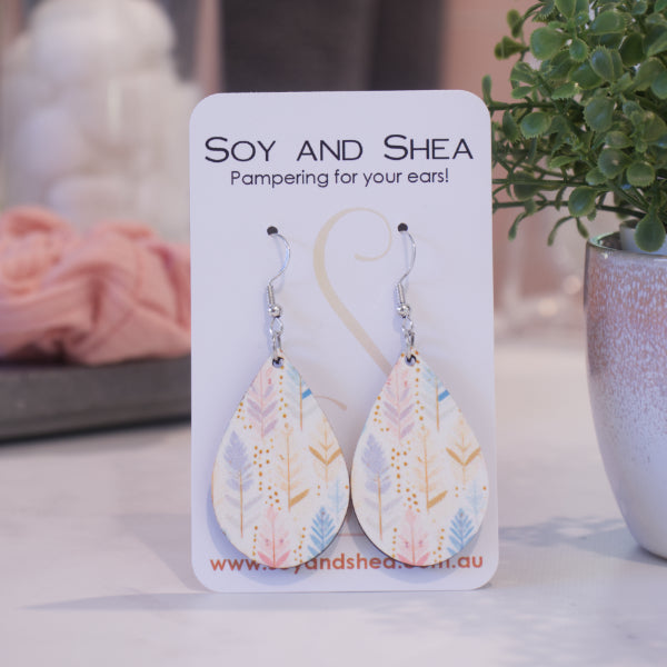 A pair of earrings on silver hooks hang on a white card branded with SOY AND SHEA leaning against a pot with a green leafy plant and a blurred background.  The earrings are teardrop shaped with boho stylised feathers in vertical lines in pale shades of pink, purple and blue with gold detail.