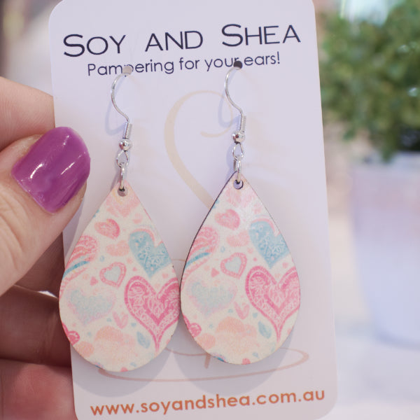 A hand holds a white card branded with SOY AND SHEA with a pair of earrings hanging on hooks.  The earrings are teardrop shaped with stylised boho hearts in pale shades of pink and blue in various sizes and randomly scattered over a white background.