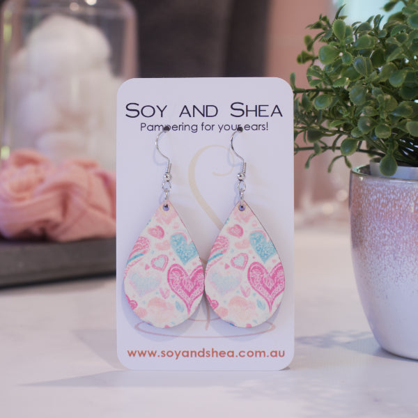 A pair of earrings on silver hooks hang on a white card branded with SOY AND SHEA leaning against a pot with a green leafy plant and a blurred background.  The earrings are teardrop shaped covered in boho stylised hearts in various shapes and sizes in bright pale blue and pink against a white background.