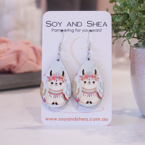 A pair of earrings on silver hooks hang on a white card branded with SOY AND SHEA leaning against a pot with a green leafy plant and a blurred background.  The earrings are teardrop shaped with an illustration of a white llama laying down surrounded by cacti and pale blue sky.  The llama has a headdress of flowers with a blanket on its back and decorations around its neck