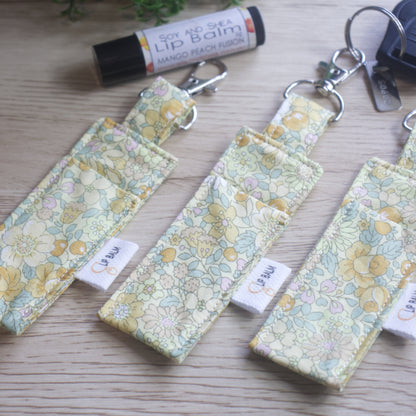A selection of three clip balms showing variations in fabric design, lie on a wooden side with the middle Clip Balm attached to a car key.  A lip balm lays near-by along with some greenery.  The Clip balms are a pale yellow colour covered in a floral design of bramble like flowers and berries in tones of deep yellow and soft green leaves.