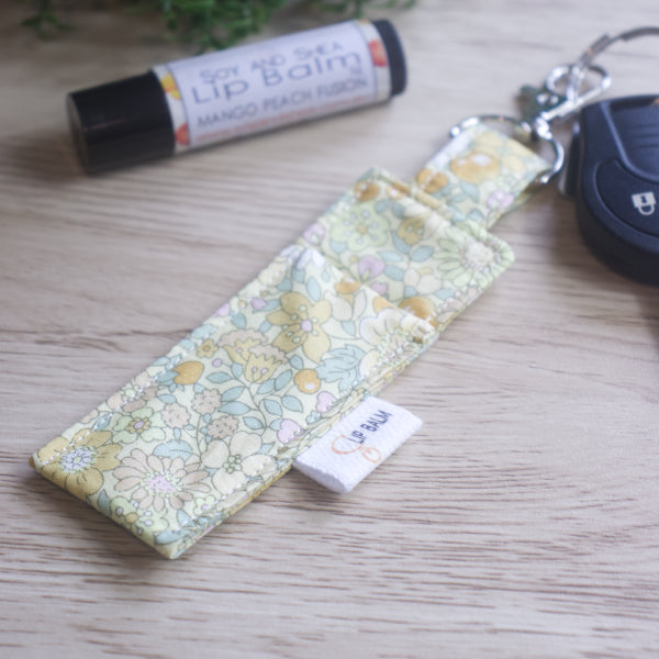 A lip balm holder lies on a wooden surface attached to a car key with a "lobster claw" clip.  To the side is a tube of lip balm. The Clip Balm has a pale yellow fabric covered in a floral design with bramble like flowers in shades of yellow and green leaves.