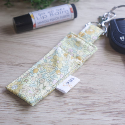A lip balm holder lies on a wooden surface attached to a car key with a "lobster claw" clip.  To the side is a tube of lip balm. The Clip Balm has a pale yellow fabric covered in a floral design with bramble like flowers in shades of yellow and green leaves.