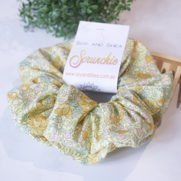 A colorful scrunchie sits on wooden surface resting against a wooden comb with greenery in the background.  A cardboard tag reads SOY AND SHEA SCRUNCHIE.    The scrunchie has a pale yellow background and is covered in bramble like flowers and berries in deeper yelllow shades with pale green leaves.