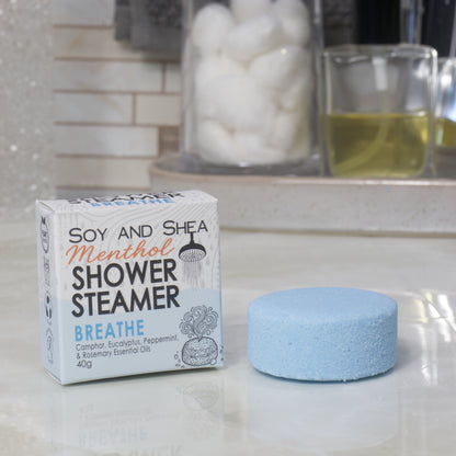 A box of Breathe Soy and Shea Shower Steamer next to a round, light blue steamer disc on a white countertop, with a blurred bathroom background showing cotton pads and a glass of oil.