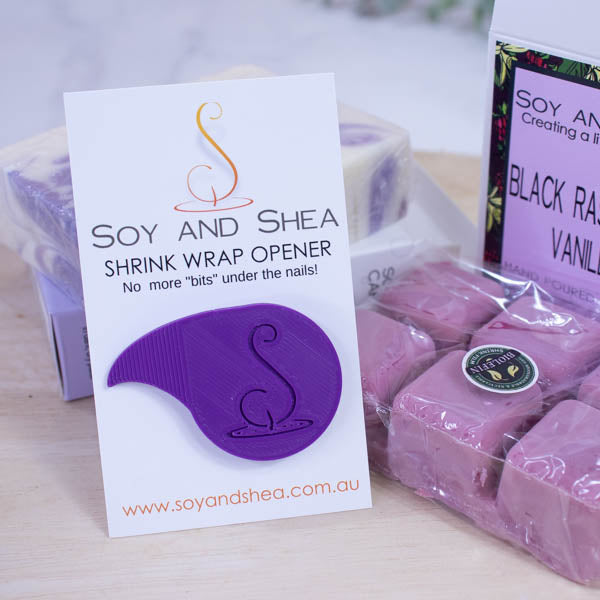 A white business-sized card branded SOY AND SHEA leans against a purple and white bar of soap and a large magenta coloured melt block both wrapped in a plastic film.   Additional text on the card reads SHRINK WRAP OPENER NO MORE BITS UNDER THE NAILS.   Stuck to the card is a teardrop shaped shrinkwrap opener that has the logomark embossed into with the point of the teardrop bent into a sharp point.  The opener is a rich bright purple.