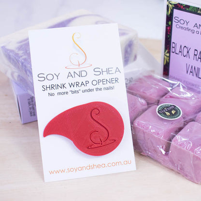 A white business-sized card branded SOY AND SHEA leans against a purple and white bar of soap and a large magenta coloured melt block both wrapped in a plastic film.   Additional text on the card reads SHRINK WRAP OPENER NO MORE BITS UNDER THE NAILS.   Stuck to the card is a teardrop shaped shrinkwrap opener that has the logomark embossed into with the point of the teardrop bent into a sharp point.  The opener is bright red