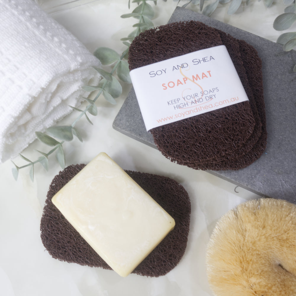 A flatlay image showing a collection of Brown Soap mats, displayed on a bathroom countertop along with a white towel, body brush and greenery.  Three soap mats are stacked upon each other in a fan shape on a grey stone tray with the top one featuring a product label.  A fourth mat lies to the bottom of the image with a cream soap lying on it.