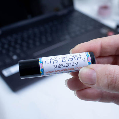 A hand holds a black tube of lip balm against a blurred background with an open laptop computer on a white surface.  The label has text LIP BALM BUBBLEGUM against a white background with a bright border in shades of blue, pink and yellow