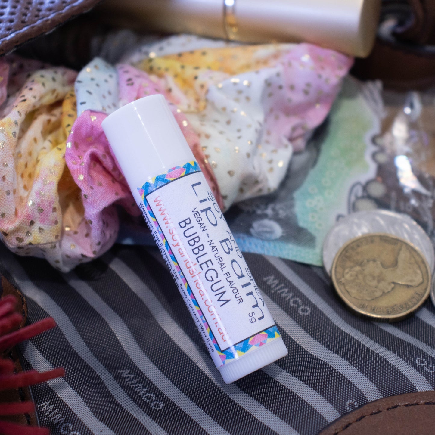 A white tube of lip balm lies on an open handbag surrouded by a scrunchie and money.  The label has a blue pink and yellow abstract design