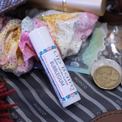 A white tube of lip balm lies on an open handbag surrouded by a scrunchie and money.  The label has a blue pink and yellow abstract design