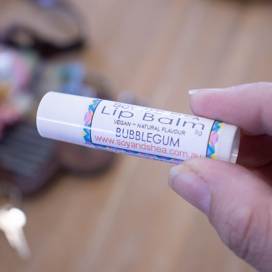 A hand holds a white tube labelled lip balm over a blurred image of an open handbag with contents spilling onto the table.  The lip balms features a label with an abstract design in blue, pink and yellow