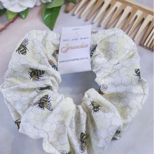 A Bumblebee Scrunchie, handcrafted from premium cotton with a charming honeycomb and bee pattern, sits elegantly on a surface. Its tag displays "Soy and Shea" along with the website "soyandshea.com.au." Nearby, a wooden comb and green leaves accentuate its appeal, highlighting its quality cotton fabric and knotless elastic design.