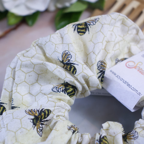 Close-up of the Bumblebee Scrunchie by Soy and Shea, showcasing its handmade design with a bee and honeycomb pattern. Made from premium cotton fabric, this scrunchie features a knotless elastic for added comfort. A small fabric tag with a partially visible logo is attached. The soft focus background includes greenery and textured elements.